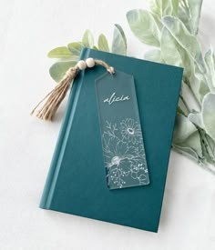 a book with a tag attached to it sitting next to some leaves and flowers on a white surface