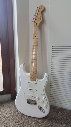 a white electric guitar sitting on the floor next to a wall with a window behind it