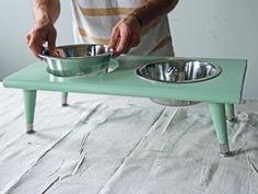 a man standing over two metal bowls on top of a green table next to a white wall