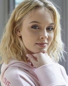 a woman with blonde hair and blue eyes wearing a pink sweatshirt looking at the camera