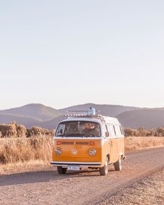 an orange vw bus driving down a dirt road in front of some grass and mountains