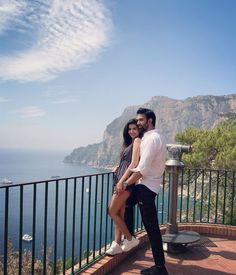 a man and woman standing next to each other on a balcony overlooking the ocean with mountains in the background