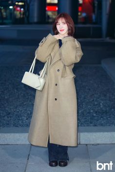 a woman is standing on the sidewalk wearing a coat and holding a purse in her hand