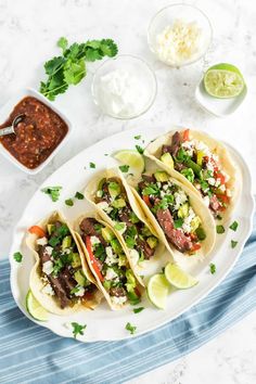 three steak tacos on a plate with salsa and limes