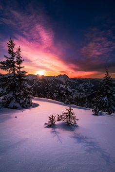 the sun is setting over some snow covered hills and trees in the foreground, with mountains in the background