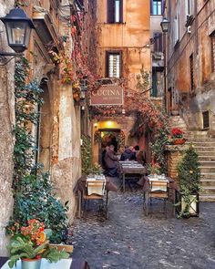 two people sitting at an outdoor table in the middle of a narrow alleyway between buildings