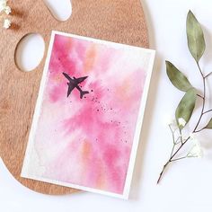 a card with an airplane painted on it next to flowers and a paintbrush in the background