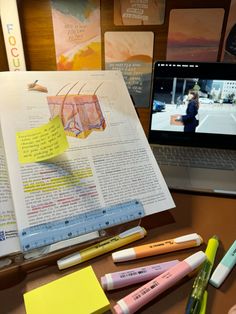 an open book sitting on top of a desk next to a laptop computer and various pens