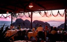 people are sitting at tables in front of an ocean view restaurant with the sun setting