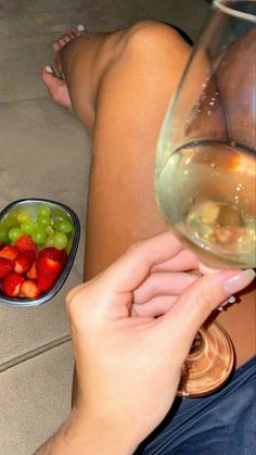 a person holding a wine glass next to a bowl of grapes and strawberries on the floor