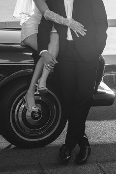 a man and woman standing next to each other in front of a car with their hands on the wheel