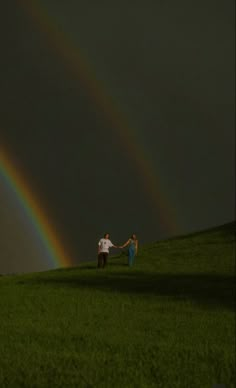 two people holding hands while standing in the grass with a rainbow in the sky behind them