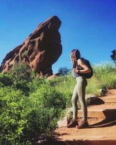 a woman is standing on some steps in front of large rocks and green grass with her back to the camera