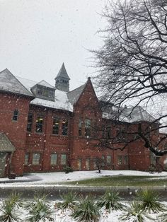 an old red brick building with snow falling on the ground and trees in front of it