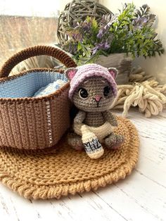 a small crocheted cat sitting next to a basket on a mat with flowers in the background