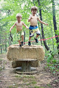 two young boys jumping over a hay bale in the middle of a wooded area