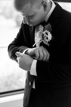 a man in a tuxedo is looking down at his watch while standing by a window