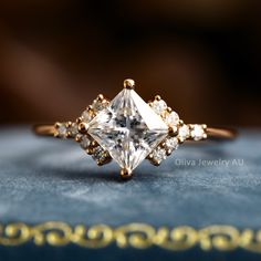 a close up view of a ring with a princess cut diamond in the center on a blue velvet surface