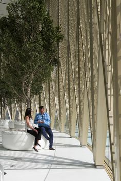 two people sitting on a bench in front of a wall with trees and glass walls