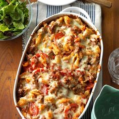 a casserole dish with meat, cheese and vegetables on a table next to a salad