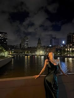 a woman in a black dress is standing on a ledge by the water at night