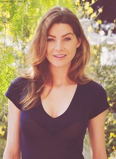 a woman standing in front of some trees and bushes smiling at the camera with her hands on her hips