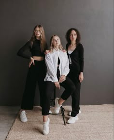 three women standing next to each other in front of a gray wall with one woman sitting on a chair