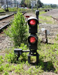 two traffic lights sitting next to each other on the side of train tracks near grass and bushes