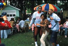 three people are standing under an umbrella in the grass with other people sitting and standing around