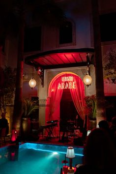 people are sitting at tables in front of a pool with red curtains and lights on it