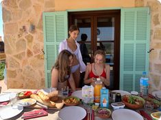 three women sitting at a table with food and drinks in front of the door to a house