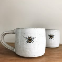 two white mugs with bees on them sitting on a wooden table next to each other