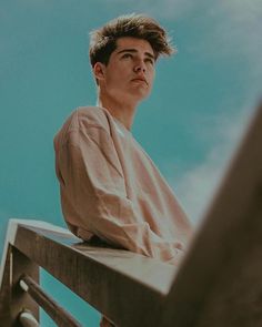 a young man sitting on top of a wooden bench next to a blue cloudy sky