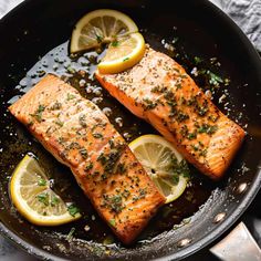 two salmon fillets in a skillet with lemons and herbs