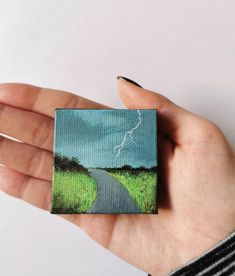 a hand holding a miniature painting of a road and lightning in the sky above it