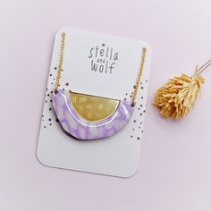 a purple and gold necklace sitting on top of a white card next to a dried flower