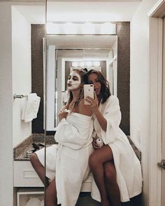 two women in white robes are taking a selfie while sitting on a bathroom counter