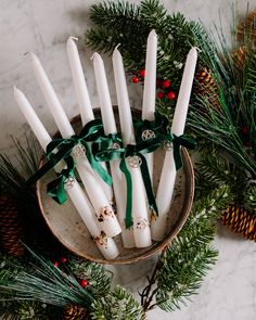 white candles tied with green ribbon in a bowl surrounded by pine cones and evergreen branches