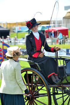 two women dressed in period clothing and hats riding on a horse drawn carriage at an event