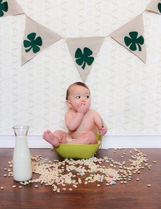 a baby sitting in a green bowl next to a milk jug and shamrock bunting