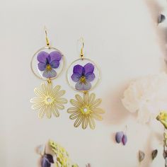 two purple flowers are hanging from gold hoops on a white surface with dried flowers in the background