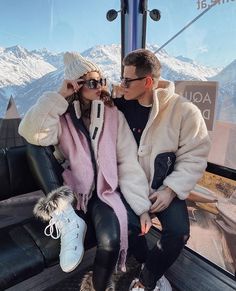 a man and woman sitting on top of a ski lift with mountains in the background