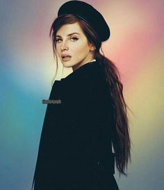 a woman with long hair wearing a black coat and hat standing in front of a colorful background