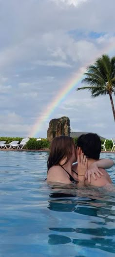 two people in a swimming pool with a rainbow in the sky and palm trees behind them