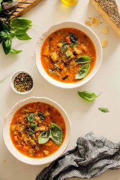 two white bowls filled with soup next to bread and basil leaves on a table top