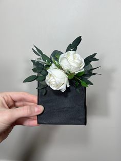 a hand holding a black bag with white flowers in it and greenery on top