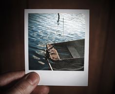 a person holding up a polaroid photo of a boat in the water with a fishing pole sticking out of it