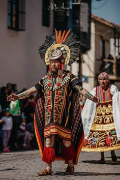 two people dressed in costumes and headdress walking down the street with their arms outstretched