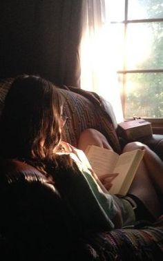 a woman sitting on top of a couch reading a book next to a window with the sun shining through