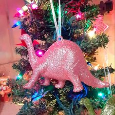 a pink dinosaur ornament hanging from a christmas tree decorated with lights and ornaments